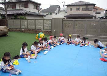 園庭に広げたシートの上に座りi一列になってお弁当を食べる園児たち