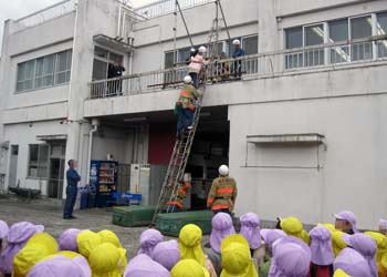 救助訓練で、消防署２階のテラスより梯子をつかって消防士に救助される保育士の様子を見守る子ども達。